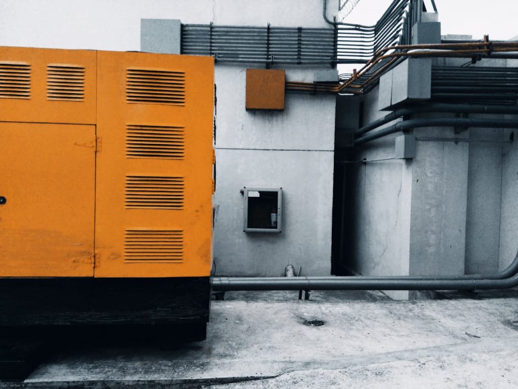 Outdoor view of an industrial generator with visible piping and wiring against a concrete wall.
